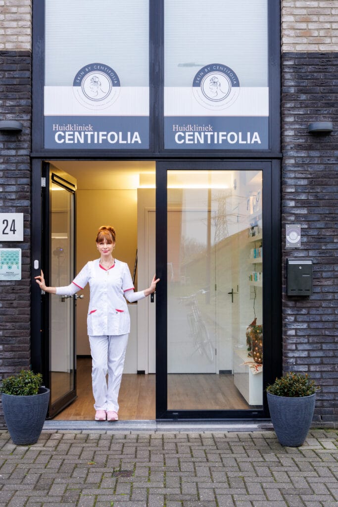 Huidkliniek Centifolia ingang met medisch professional in wit uniform bij de deuropening van moderne huidverzorgingskliniek met huisnummer 24 en sierplanten